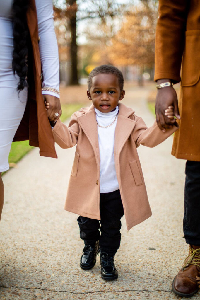 young stylish black boy holding parents hands wearing a trench coat