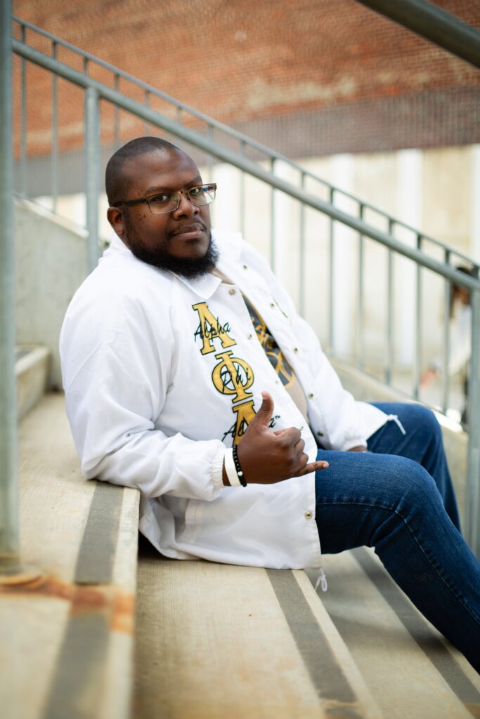 grad photos in alpha phi alpha fraternity jacket at raleigh union station man sitting on stairs doing fraternity hand sign