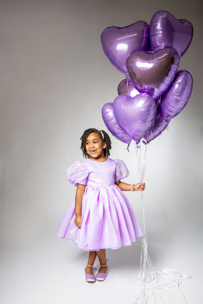 Kennedy Grace holding purple heart-shaped balloons in a purple dress during her 5th birthday photoshoot on a white backdrop.