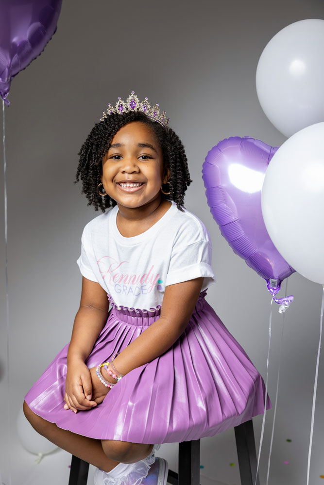 Kennedy posing on a white stool in a purple leather pleated skirt and personalized t-shirt during her birthday photoshoot.