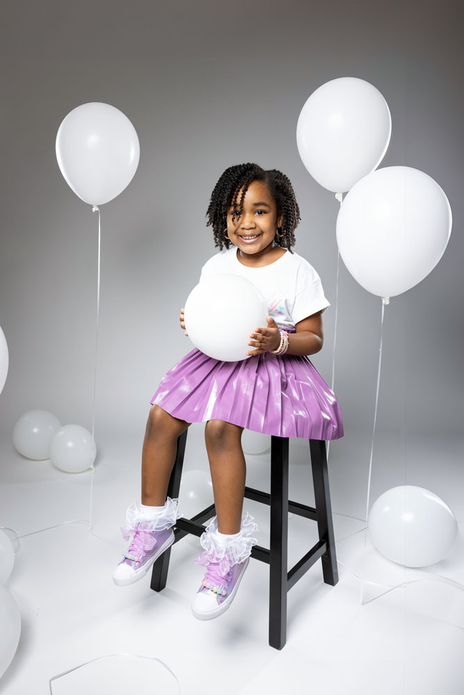 Kennedy sitting on a stool holding a white balloon in her purple leather pleated skirt and personalized t-shirt.