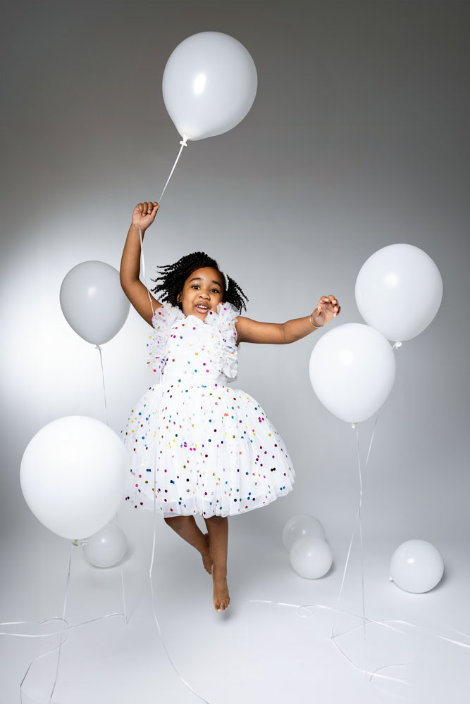 Kennedy jumping in a white dress with confetti print, holding a white balloon during her 5th birthday photoshoot.