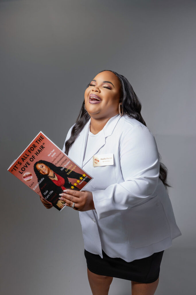 Madilla in her stylist smock, laughing and holding a branded magazine highlighting her brand's success.