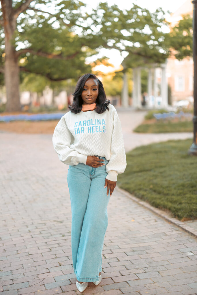 Black woman wearing a Carolina Tar Heels sweater posing on campus walkway during UNC Chapel Hill grad photos.