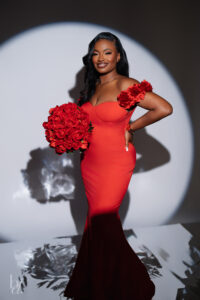 Woman standing in a red off-the-shoulder dress holding a bouquet of red roses inside a spotlight at a Raleigh birthday photoshoot.