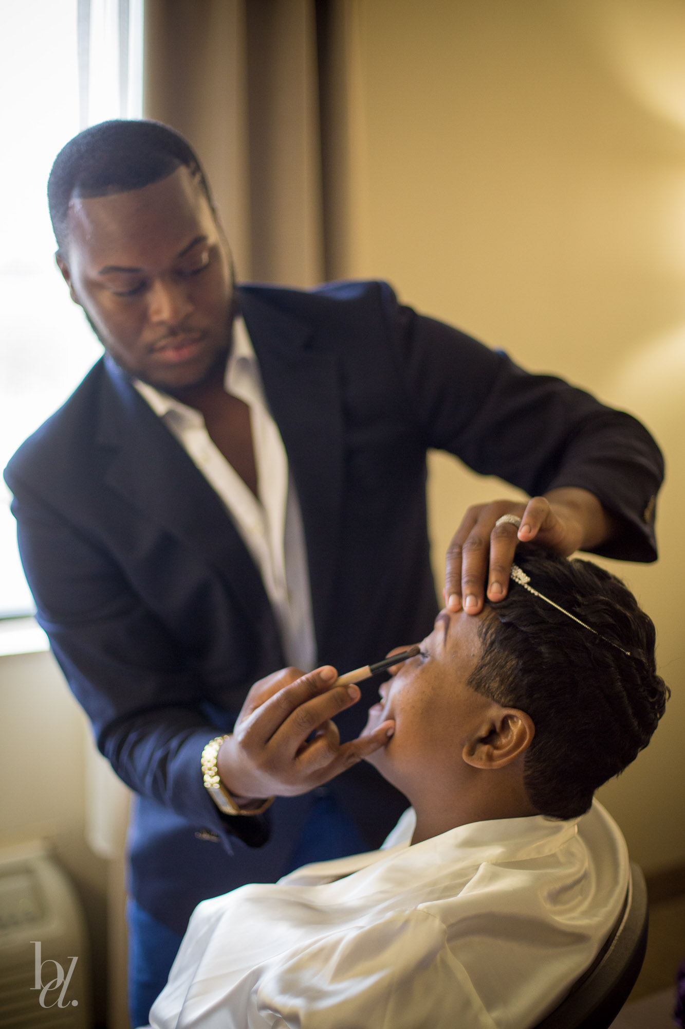 Photoshoot makeup prep as a makeup artist applies eye makeup to a client in a white robe near a window.