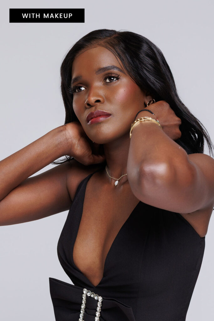 Studio portrait of a woman in a black dress with soft glam makeup, looking up with her hands near her hair.