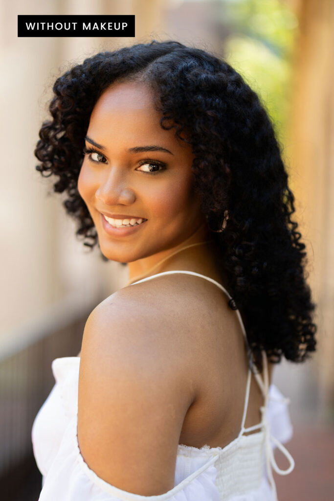 Outdoor portrait of a smiling woman with curly hair wearing a white dress, shown without makeup in warm natural light.