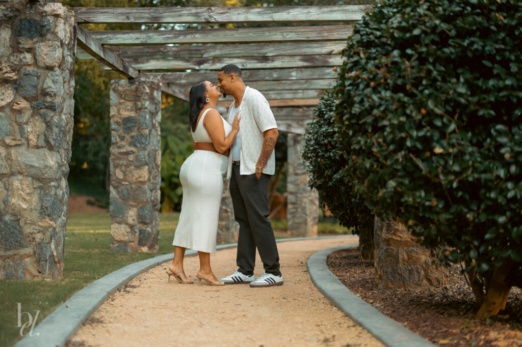 Couple standing closely under a stone pergola during an engagement photo session at the Raleigh Rose Garden.