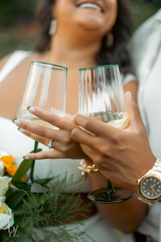 Close-up of couple holding champagne glasses and showing engagement ring during a picnic moment at an engagement photoshoot in Raleigh.