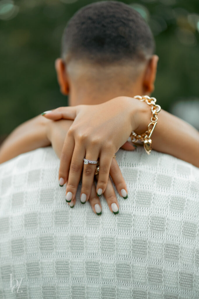 Detail shot of hands wrapped around a partner’s shoulders highlighting an engagement ring during a garden session in Raleigh.
