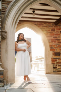 Doctoral graduate posing under Duke Chapel archway in white dress during Duke doctoral grad photos