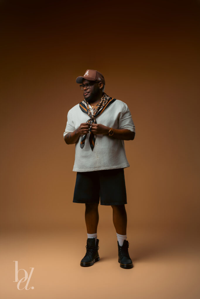 Man standing in a photography studio wearing a cream textured shirt, black shorts, brown baseball cap, black boots, and a leopard print scarf while adjusting the scarf against a warm brown seamless backdrop.