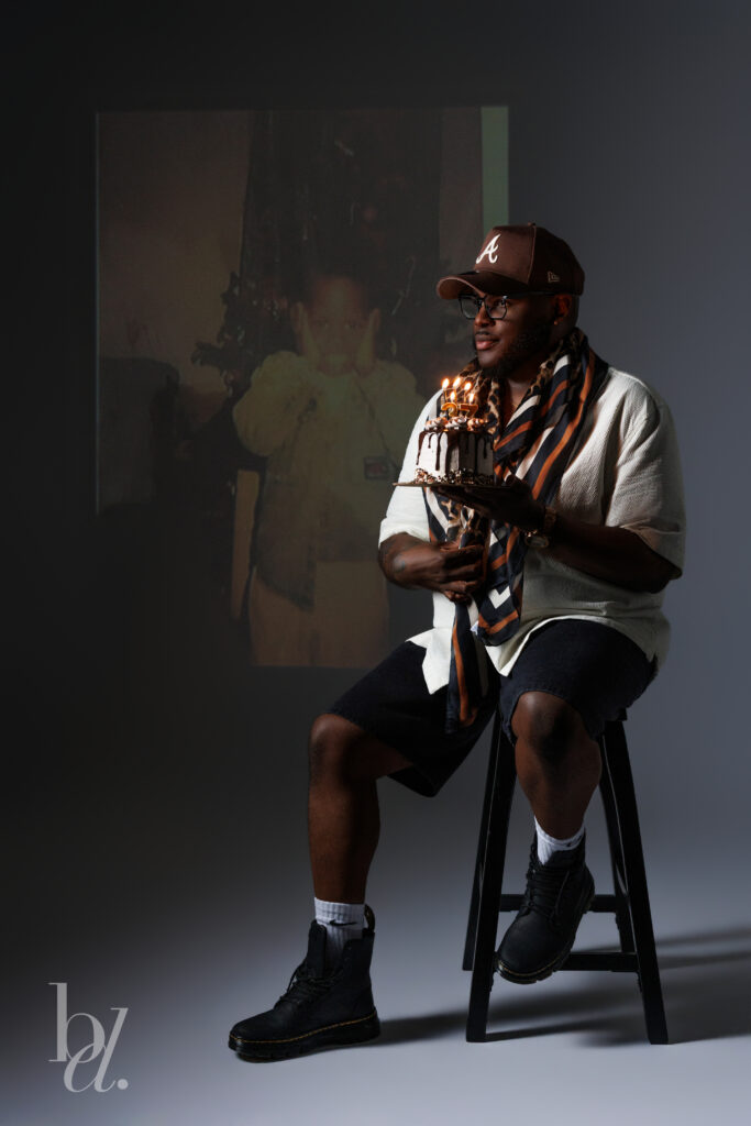 Man seated on a stool holding a birthday cake with lit candles while a childhood photograph is projected behind him, creating a nostalgic editorial birthday portrait about reflection and growth.