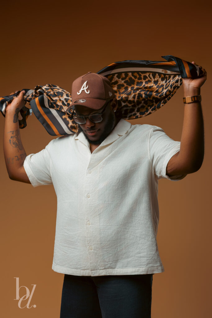 Close-up portrait of a man lifting a leopard print silk scarf over his shoulders while wearing a cream short-sleeve button-up shirt, brown baseball cap, glasses, and gold watch in a warm-toned studio.