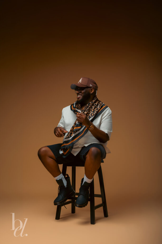 Editorial portrait of a man sitting on a black stool smiling off camera while wearing a cream textured shirt, leopard scarf, brown baseball cap, and black boots in a minimalist studio.