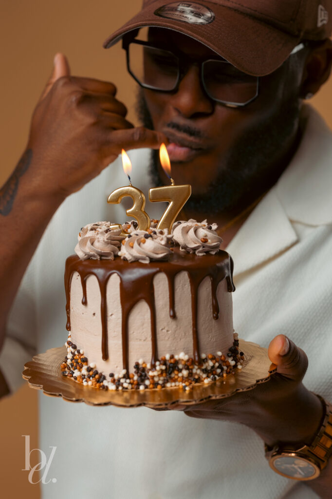 Close-up portrait of a man holding a chocolate drip birthday cake with gold number 37 candles while making a playful hand gesture before blowing out the candles.