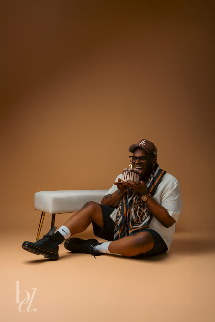 Man sitting on the studio floor laughing while holding a chocolate birthday cake with gold 37 candles beside a white upholstered bench against a warm brown backdrop.