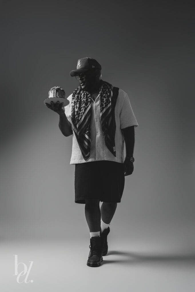 Black and white full-length portrait of a man walking through a photography studio while holding a birthday cake and wearing a cream shirt, patterned scarf, black shorts, and combat boots.