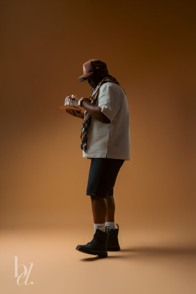 Side profile portrait of a man holding a chocolate birthday cake while looking down at the candles, photographed against a warm brown seamless background.