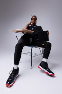 Graduate sitting in a classroom desk holding a graduation cap during a creative studio session.